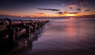 a long old rusty pipe stretches out from the beach in to the sea at sunrise