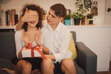 Young man giving gift and clothing eyes of girlfriend at home, woman with white package with red bow in cozy room.
