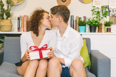 Young man giving gift and kissing girlfriend at home, attractive woman holding white package with red bow in cozy room.