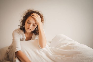 Sad young woman sitting on bed, feeling upset with hand on head at home.