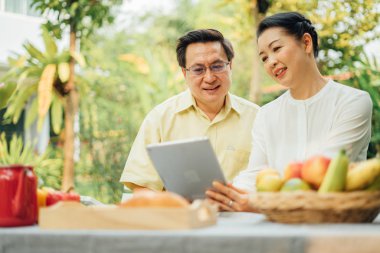 Rahatlarken ve dışarıda piknik yaparken dijital tablet kullanan mutlu Asyalı yaşlı çift portresi.