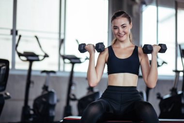 Salondaki genç ve güzel kız halterlerle egzersiz yapıyor ve gülümsüyor. Genç atletik kız halterlerle egzersiz yapıyor. Spor salonunda spor salonunda spor salonunda spor salonunda spor yapan kaslı bir kadın..