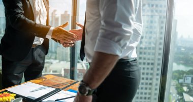 İki iş adamı pencerenin kenarında otururken el sıkışıyor. Olgun iş adamı, şehre tepeden bakan bir ofis kulesinde, modern bir iş yerindeki bir meslektaşıyla bilgi tartışıyor..