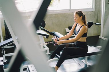 Genç fitness kızı spor salonundaki simülatörde asansörleri kaldırıyor. Spor salonundaki spor aletleriyle çalışan insanlar..