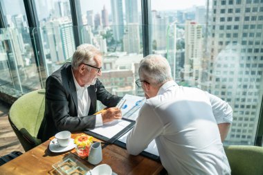 İki iş adamı pencerenin kenarında otururken el sıkışıyor. Olgun iş adamı, şehre tepeden bakan bir ofis kulesinde, modern bir iş yerindeki bir meslektaşıyla bilgi tartışıyor..