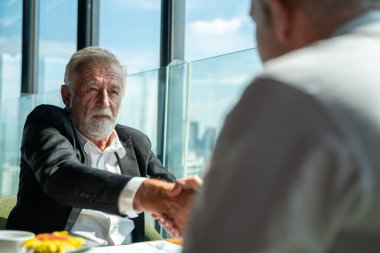 İki iş adamı pencerenin kenarında otururken el sıkışıyor. Olgun iş adamı, şehre tepeden bakan bir ofis kulesinde, modern bir iş yerindeki bir meslektaşıyla bilgi tartışıyor..