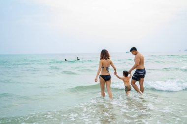 Tropikal yaz tatilinde kumsalda denizin kenarında el ele tutuşan mutlu Asyalı aile..