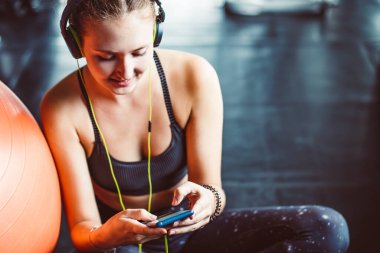 Kulaklıklı genç bir kadın spor salonunda sıkı çalıştıktan sonra müzik dinliyor. Rahat ol. Kuvvet eğitimi ve Vücut Geliştirme teması. Sıcak ve havalı. Güzel kız cep telefonuyla kayıyor. Kapat.