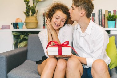 Young man giving gift and kissing surprised girlfriend at home, pleased woman looking at white package with red bow in cozy room.
