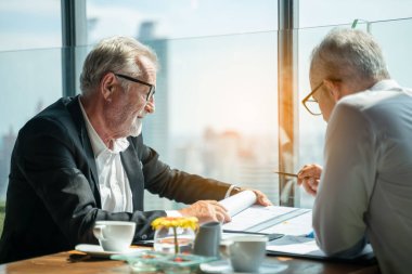 İki iş adamı pencerenin kenarında otururken el sıkışıyor. Olgun iş adamı, şehre tepeden bakan bir ofis kulesinde, modern bir iş yerindeki bir meslektaşıyla bilgi tartışıyor..