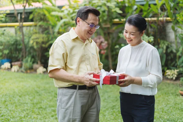 Senior Asian man showing red present box with white ribbon outdoor.Two Asian senior couple giving red present box on holiday occasion.