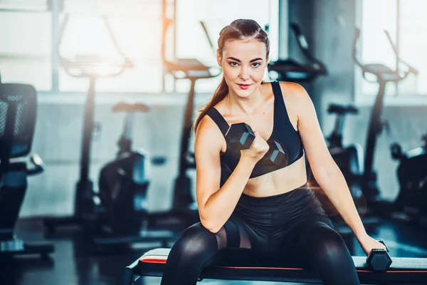 Beautiful young girl in the gym doing exercises with dumbbells and smiling.young athletics girl doing dumbbells press exercises. Fitness muscled woman in black sport clothing workout on bench in gym.