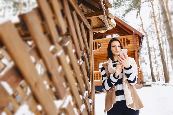 Jeune fille avec téléphone en gilet sur contre de chalet en bois — Photo