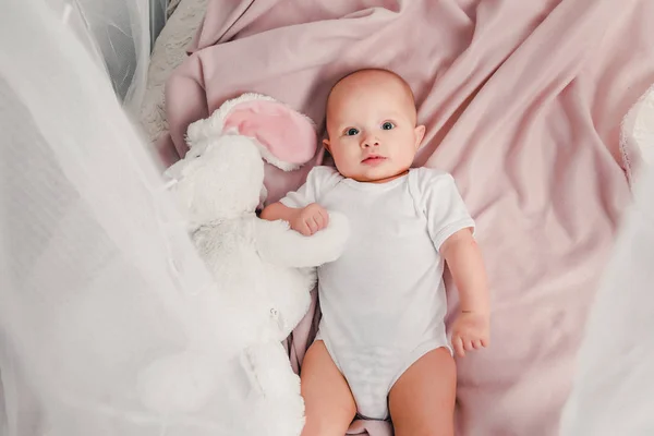 Um bebezinho deita em uma cama com um coelho de brinquedo e sorri — Fotografia de Stock