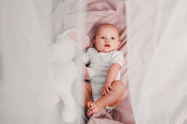 Um bebezinho deita em uma cama com um coelho de brinquedo e sorri — Fotografia de Stock