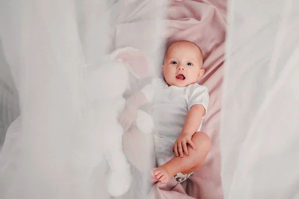 Um bebezinho deita em uma cama com um coelho de brinquedo e sorri — Fotografia de Stock