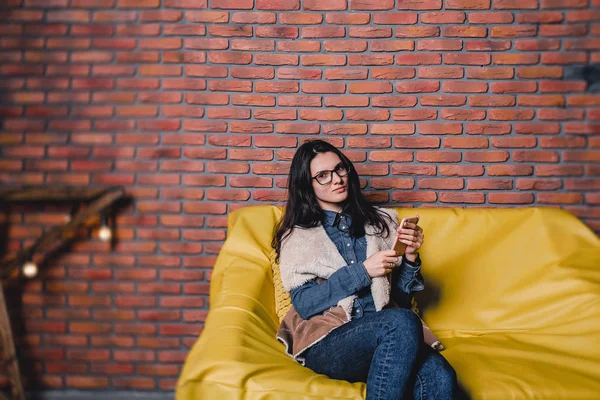 Joven bonita chica en gafas con un teléfono en un sofá contra — Foto de Stock