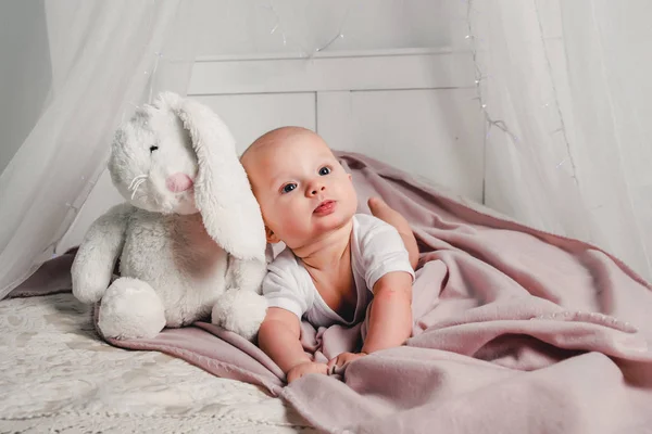 Um bebezinho deita em uma cama com um coelho de brinquedo e sorri — Fotografia de Stock