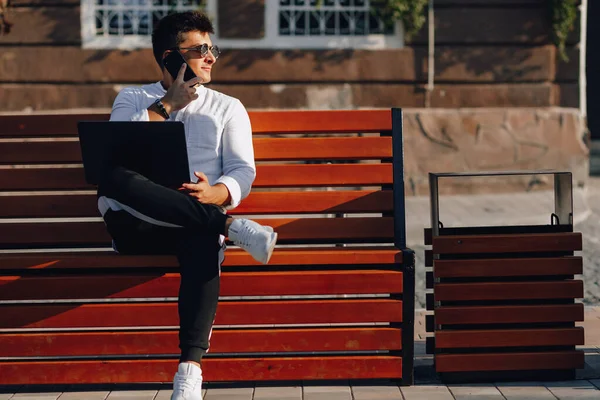 young stylish guy in shirt with phone and notebook on bench on sunny warm day outdoors, freelance