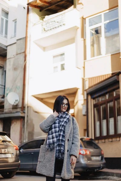 Atraente menina positiva vestindo óculos em um casaco no fundo de edifícios em carros — Fotografia de Stock