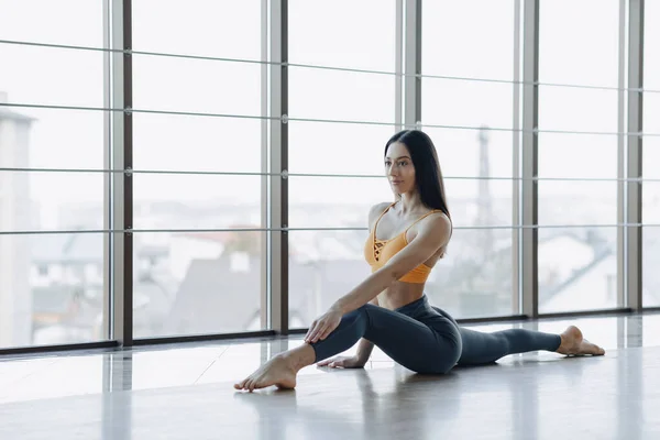Chica atractiva joven haciendo ejercicios de fitness con yoga en el suelo contra el fondo de ventanas panorámicas — Foto de Stock