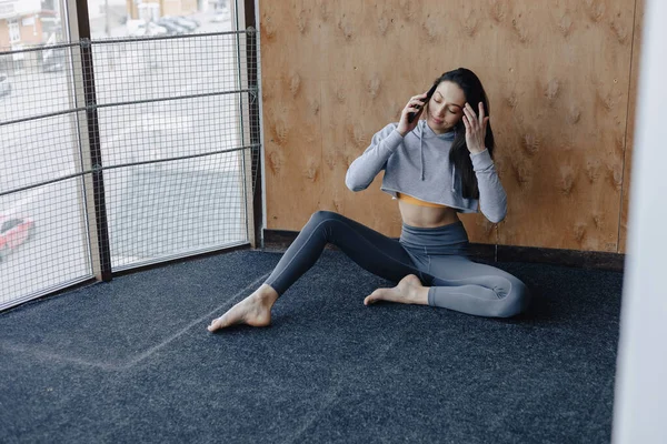 Young attractive fitness girl sitting on the floor near the window on the background of a wooden wall, resting on yoga classes and talking on phone — Stock Photo, Image
