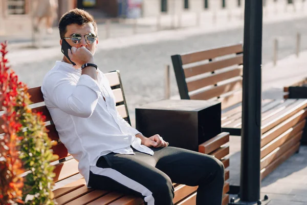 Ung snygg kille i skjorta med telefon på bänk på solig varm dag utomhus — Stockfoto