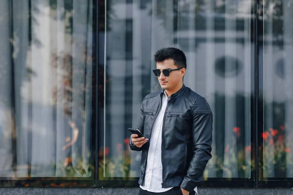 young stylish guy in glasses in black leather jacket with phone on glass background