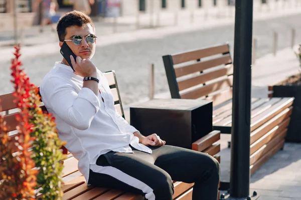 Ung snygg kille i skjorta med telefon på bänk på solig varm dag utomhus — Stockfoto