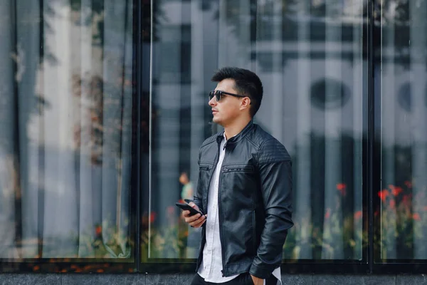 Ung snygg kille i glasögon i svart läder jacka med telefon på glas bakgrund — Stockfoto