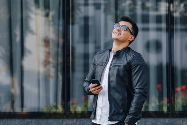 Ung snygg kille i glasögon i svart läder jacka med telefon på glas bakgrund — Stockfoto