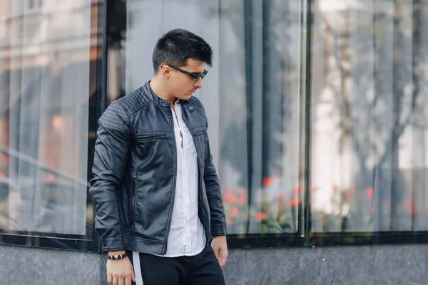 Ung snygg kille i glasögon i svart läder jacka på glas bakgrund — Stockfoto