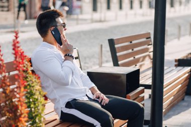 Açık havada güneşli bir günde telefonu bankta duran beyaz tişörtlü şık bir genç.