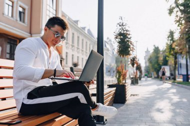 Beyaz tişörtlü, telefon ve defterli genç şık adam açık havada güneşli bir günde bankta çalışıyor. Serbest çalışıyor.