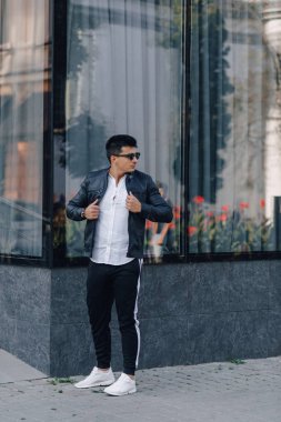 Gözlüklü, siyah deri ceketli, cam arka planda genç, şık bir adam.