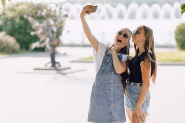 Parkta yürüyüş yapan iki güzel kız telefonda kendi resimlerini çekiyorlar. Güneşli bir yaz gününde selfieler, neşe ve dostluklar.