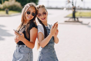 Parkta telefonla yürüyen iki güzel genç kız. Sıcak güneşli yaz günü, neşe ve dostluklar.