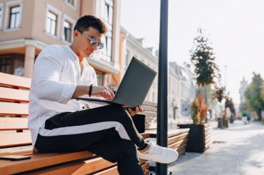 Beyaz tişörtlü, telefon ve defterli genç şık adam açık havada güneşli bir günde bankta çalışıyor. Serbest çalışıyor.