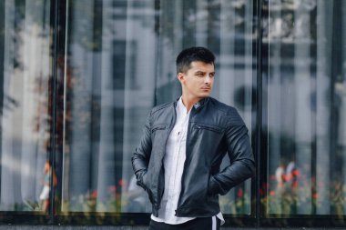 Gözlüklü, siyah deri ceketli, basit cam arka planda telefonu olan şık bir genç.