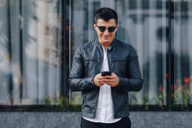 Gözlüklü, siyah deri ceketli, basit cam arka planda telefonu olan şık bir genç.