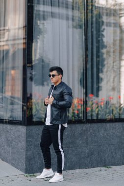 Gözlüklü, siyah deri ceketli, cam arka planda genç, şık bir adam.