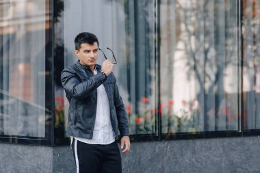 Gözlüklü, siyah deri ceketli, cam arka planda genç, şık bir adam.