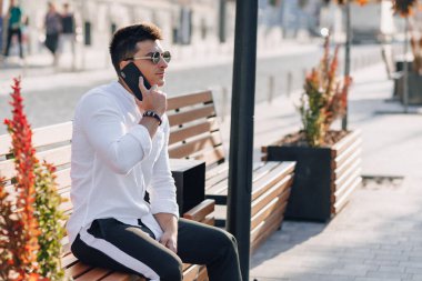 Açık havada güneşli bir günde telefonu bankta duran beyaz tişörtlü şık bir genç.