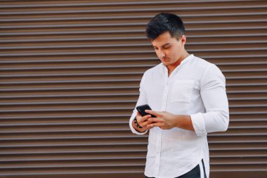 Gömlekli genç adam basit kahverengi arka planda telefonla yazı yazıyor.