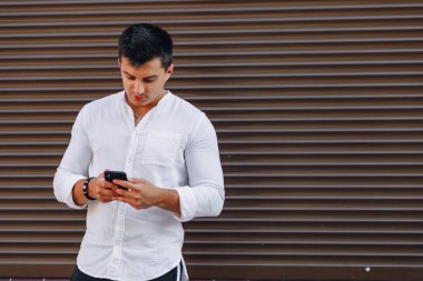 Gömlekli genç adam basit kahverengi arka planda telefonla yazı yazıyor.