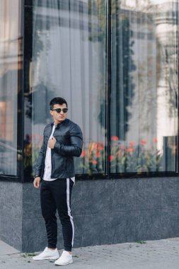 Gözlüklü, siyah deri ceketli, cam arka planda genç, şık bir adam.
