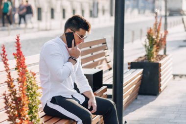 Açık havada güneşli bir günde telefonu bankta duran beyaz tişörtlü şık bir genç.