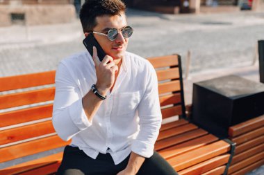 Açık havada güneşli bir günde telefonu bankta duran beyaz tişörtlü şık bir genç.