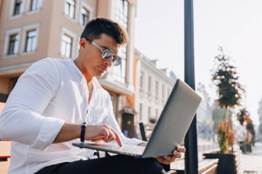 Beyaz tişörtlü, telefon ve defterli genç şık adam açık havada güneşli bir günde bankta çalışıyor. Serbest çalışıyor.