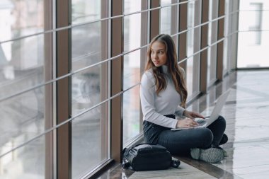 Bilgisayarla çalışan çekici bir kız havaalanı terminalinde ya da ofisin zemininde çalışıyor. seyahat atmosferi ya da alternatif çalışma ortamı. Serbest çalışan öğrenci iş toplantısına gidiyor. Alternatif iş yeri, teknoloji ve bekleme kavramı.